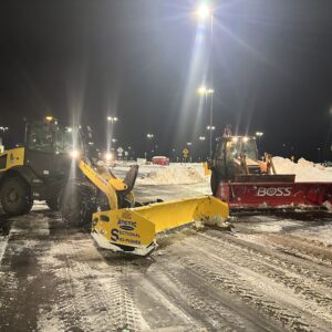 Snow removal truck operating during nighttime snowfall, plowing and de-icing large commercial parking area efficiently.