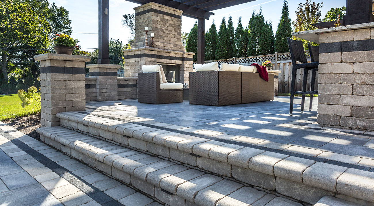 Elevated stone patio with wide steps, wicker seating, fireplace, and pergola, surrounded by trees and manicured lawn.