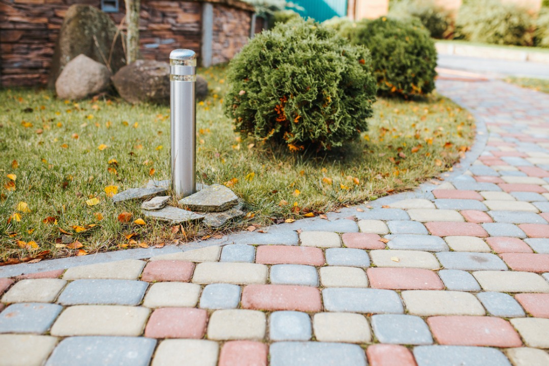 A colorful brick pathway curves through a landscaped yard with bushes, a modern bollard light, and scattered autumn leaves.