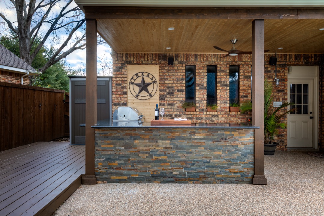 Covered outdoor kitchen with stone bar, stainless grill, and rustic decor on a patio with stamped concrete flooring.