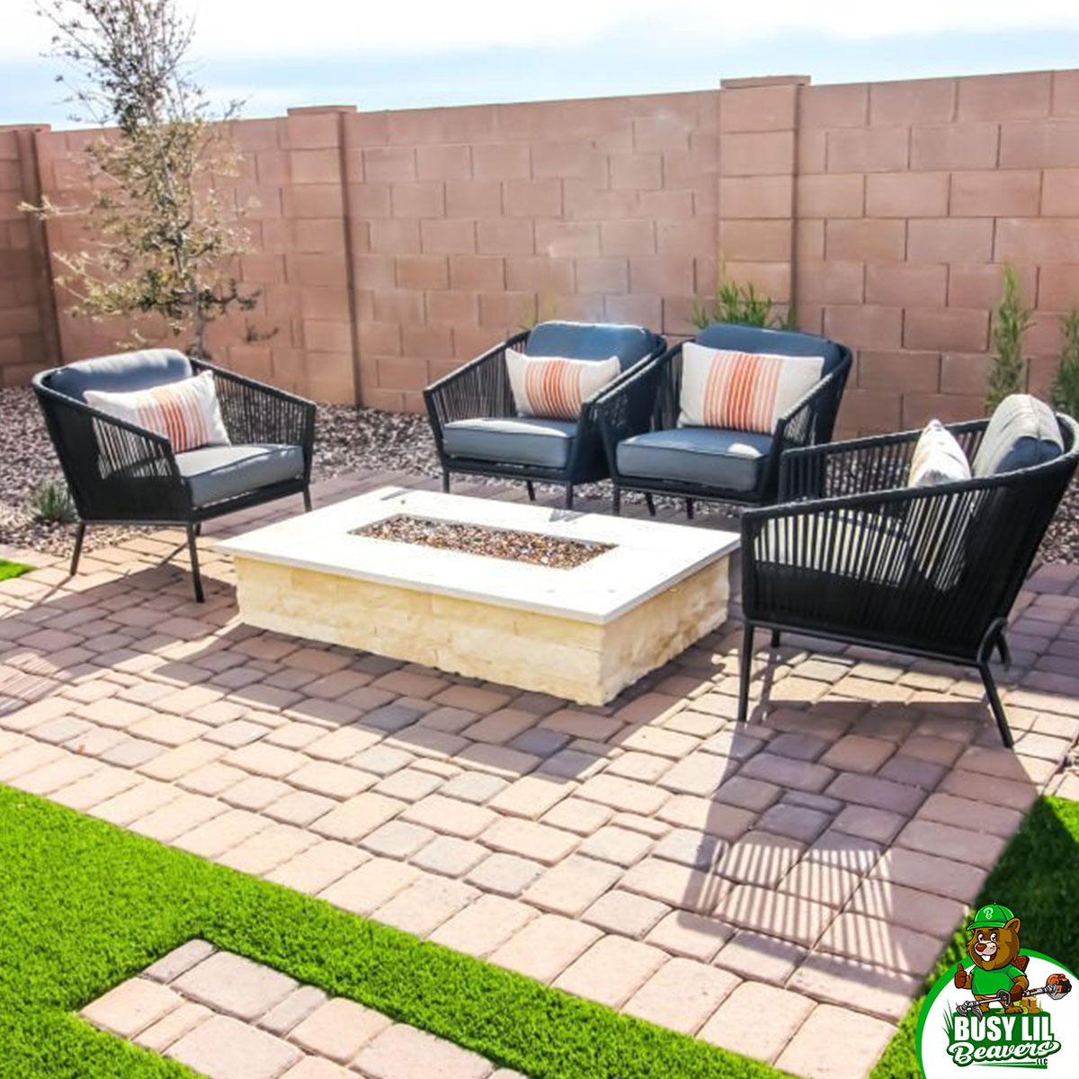 Modern patio with four cushioned chairs around a stone firepit on tan pavers, framed by artificial turf and block fencing.