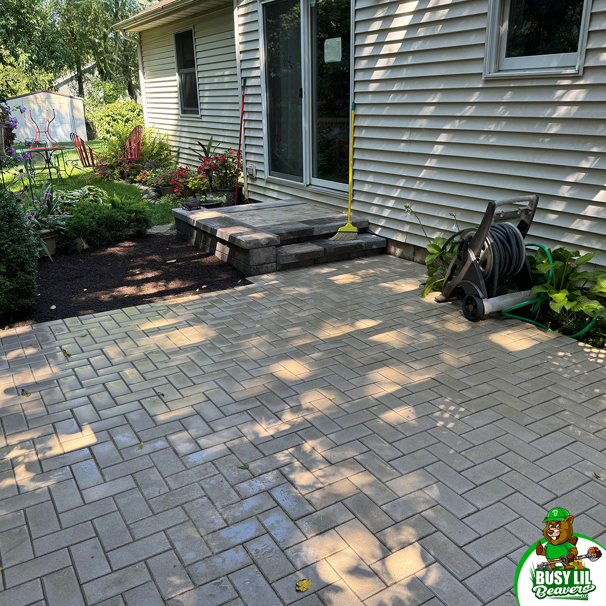 Freshly installed paver patio with small steps, hose reel, and vibrant landscaping outside a beige vinyl-sided home.