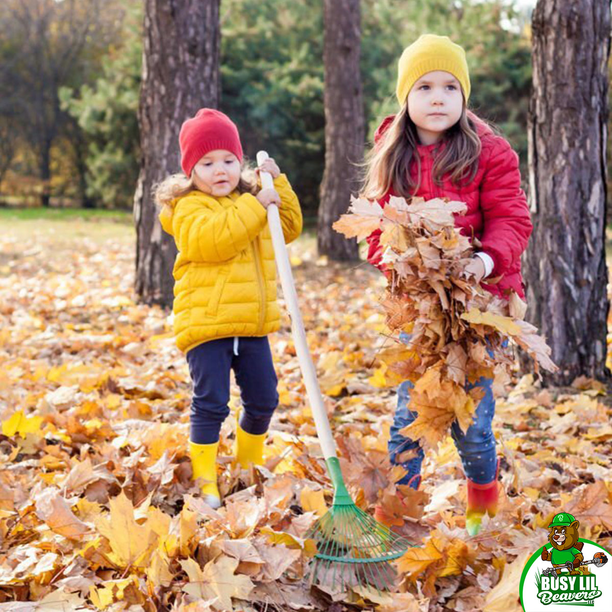 Two young children in colorful jackets and beanies rake and gather fall leaves in a forested area during autumn.