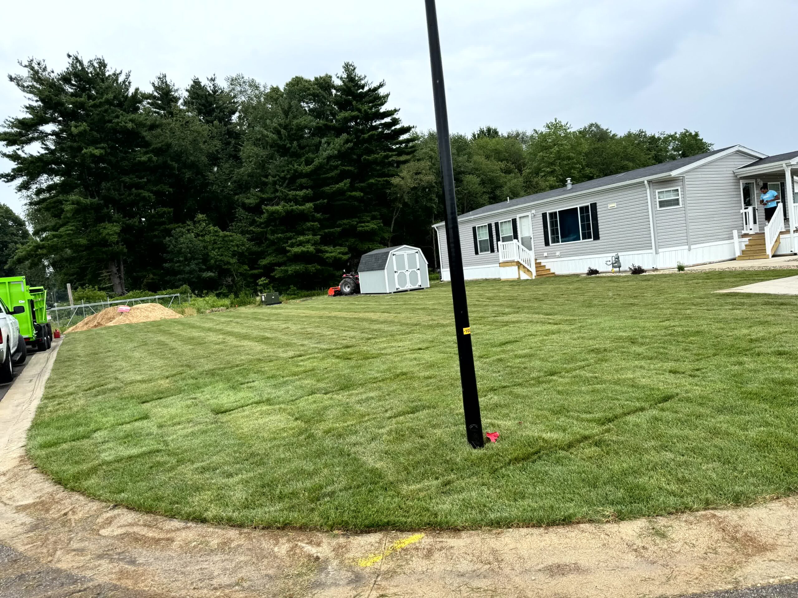 Newly installed sod covers a large lawn beside a mobile home with landscaping trucks and trees visible in the background.