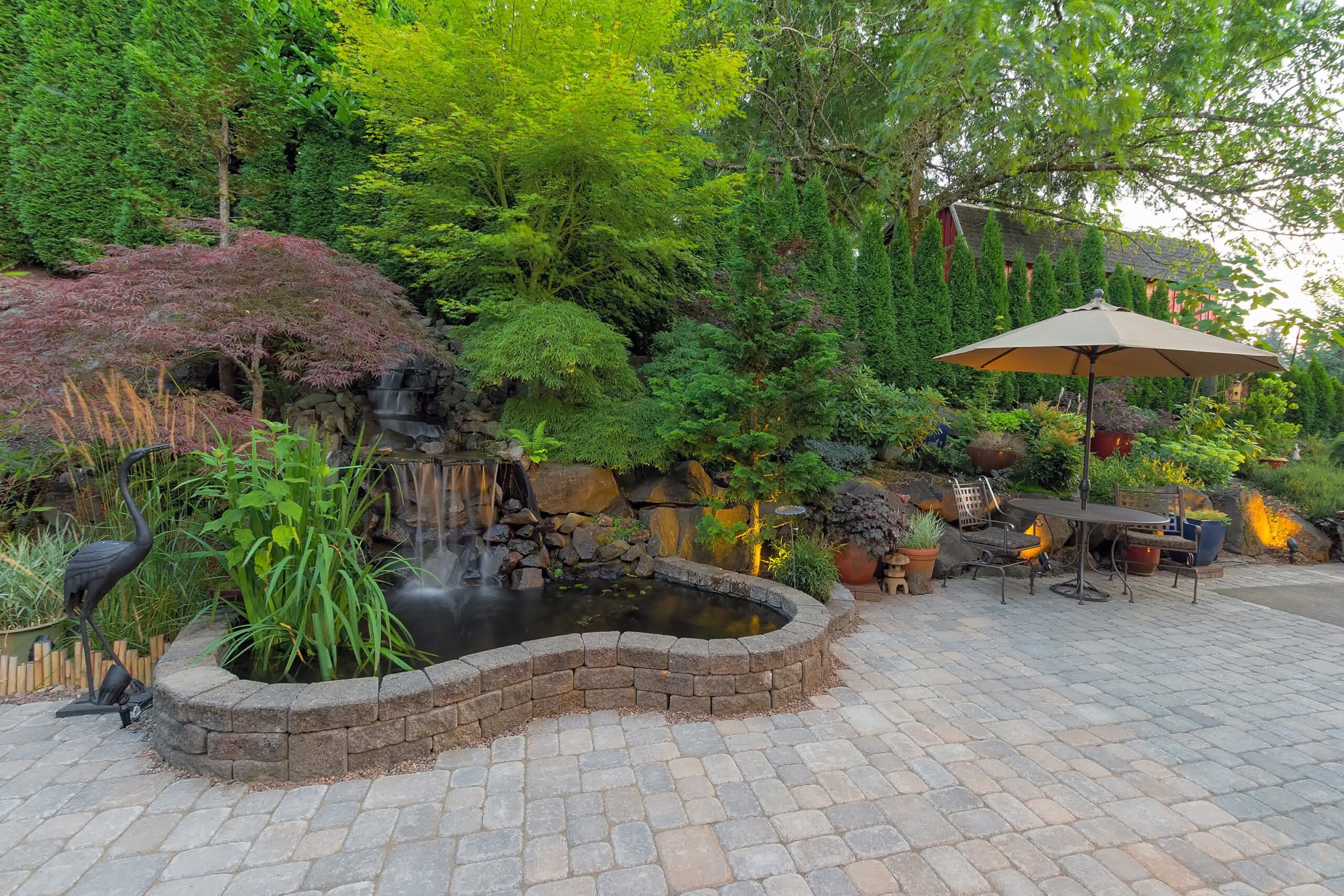 Backyard patio with paver stones, water feature, small pond, cascading waterfall, lush plants, outdoor furniture, and uplighting.