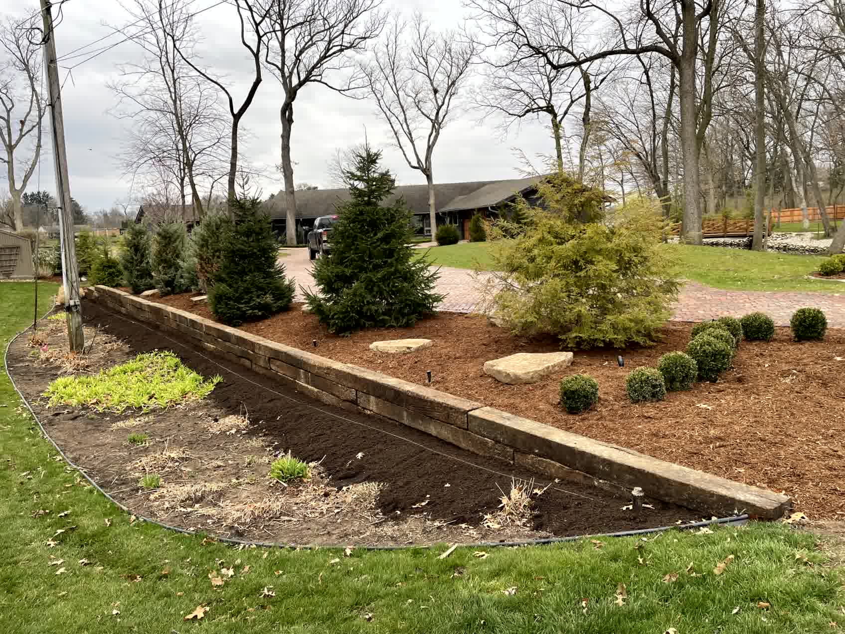 Low retaining wall made of weathered stone blocks supports mulch landscaping with evergreens along a curved residential driveway.