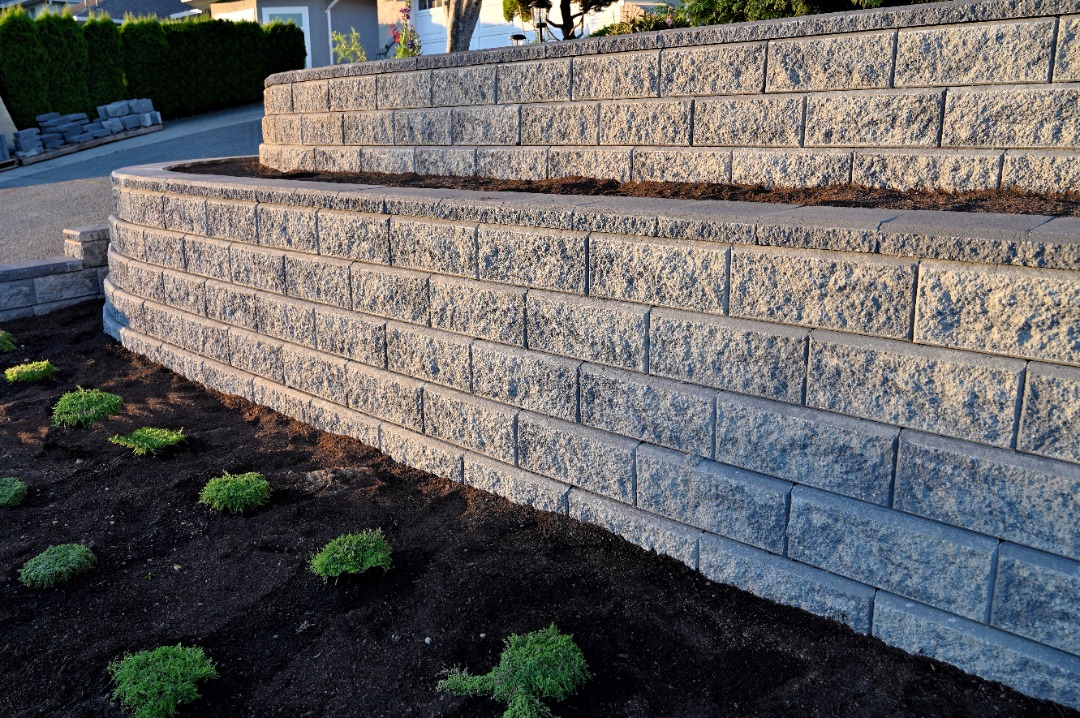 Tiered retaining wall built with gray interlocking concrete blocks, bordered by mulch beds and freshly planted ground cover.