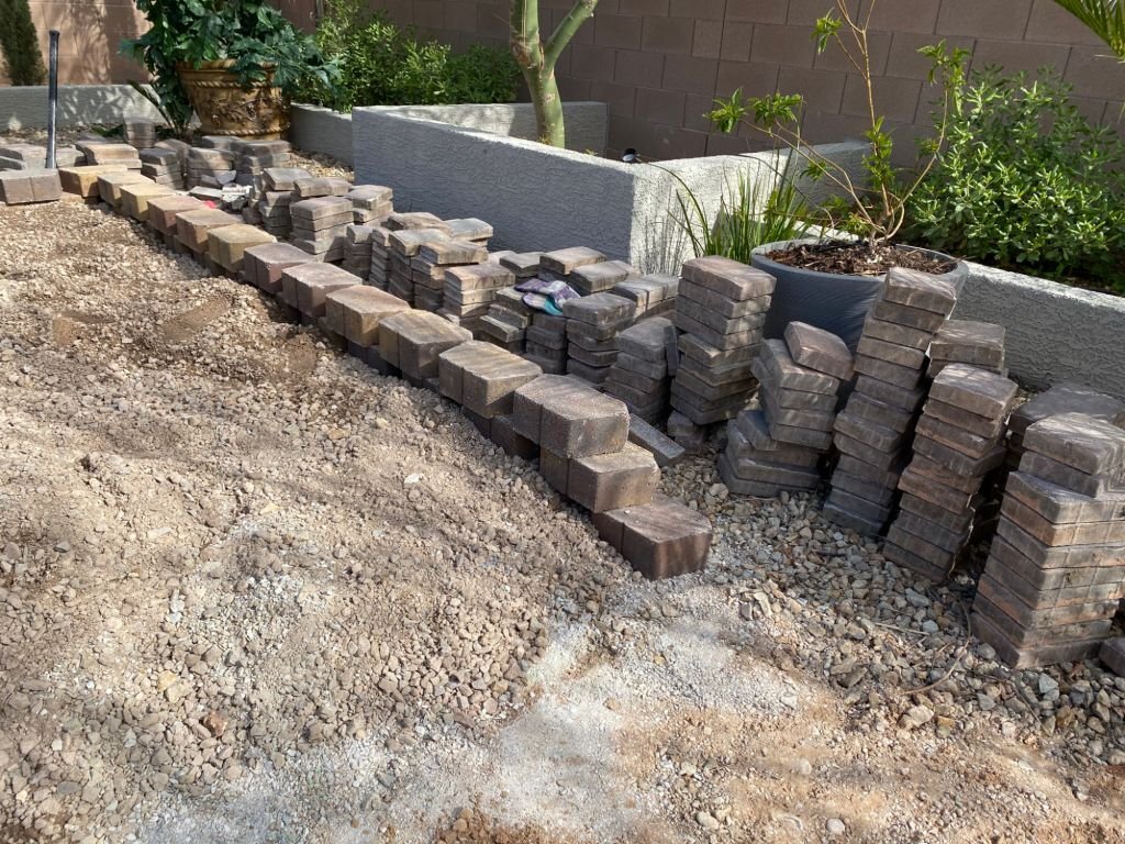 Stacks of concrete pavers organized along a dirt and gravel base, prepared for installation of a new landscape retaining wall.
